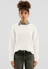 Biały sweter o strukturze waflowej z prążkowanymi mankietami, noszony na szarej koszuli w kratę. Sweter ma luźny krój i okrągły dekolt. Na dole ciemne dżinsy.