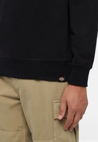 Felpa nera con polsini e orlo a coste, caratterizzata da una piccola etichetta circolare logo in basso a sinistra. Abbinata a pantaloni cargo color khaki.