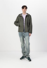 Olivengrønn hettejakke med glidelås foran, brukt over en lys lilla t-skjorte. Lyseblå jeans og mørkegrønne sneakers fullfører antrekket.