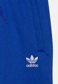 Blå mjukisbyxor i ett mjukt material med en texturerad yta, som har en vit broderad Adidas-logotyp på vänster sida nära fickan.