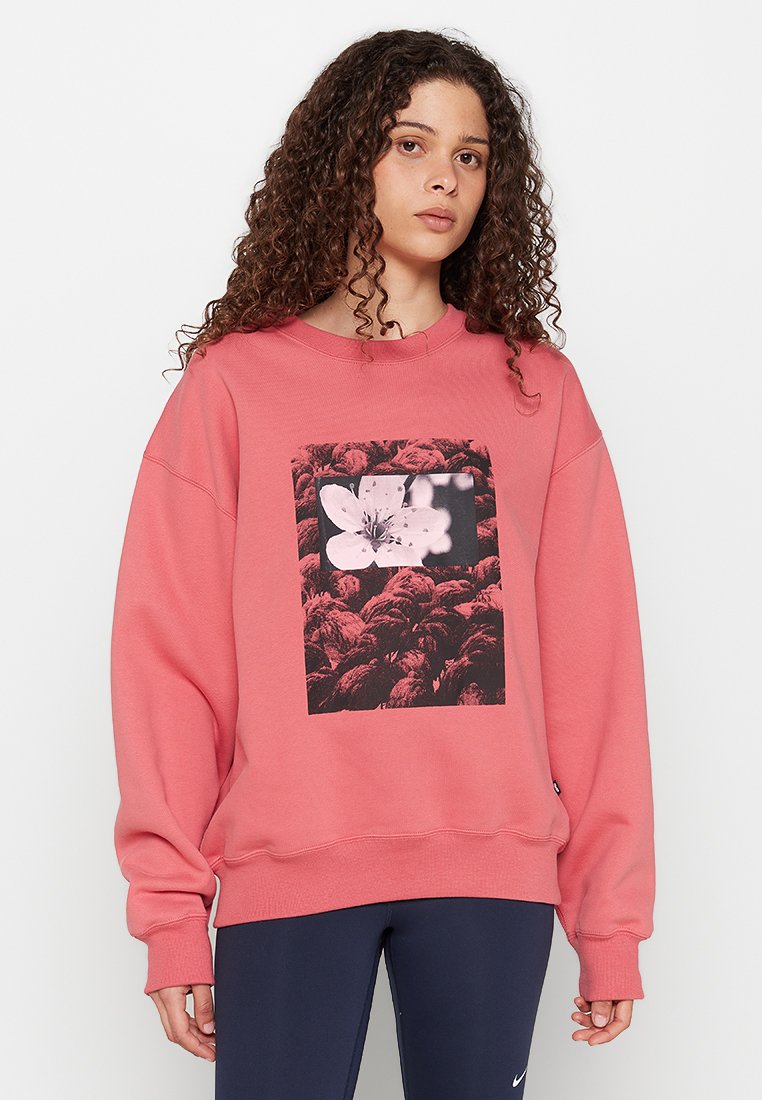 Kobieta z kręconymi włosami, ubrana w koralowo-różowy sweter z czarno-białym graficznym wzorem kwiatowym oraz granatowe legginsy.