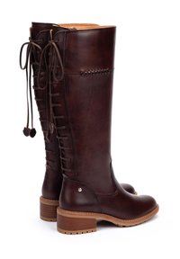 Botas de cuero marrón oscuro hasta la rodilla con cordones en la parte posterior, tacones de madera apilados y detalles de costura a lo largo de los lados exteriores.