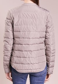 Ljusgrå puffjacka med kviltad textur, ribbad krage och synlig sömnad. Bakomvy visar en figurnära design och en lätt böjd fåll.