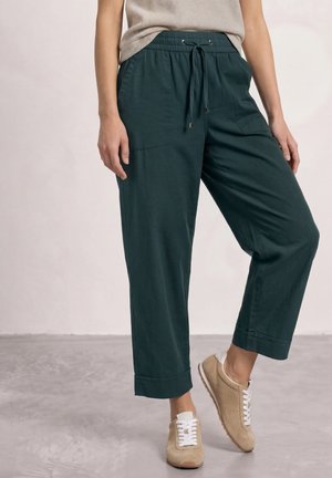 Personne portant un pantalon court vert foncé avec une taille à cordon et des baskets beiges, debout sur un sol gris sur fond uni.