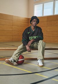 Čierny dres Nike Celtics s zelenými akcentmi, khaki nohavice s detailmi na zacvakávanie a biele tenisky s čiernym swooshom, sediaci na drevenej lavici.
