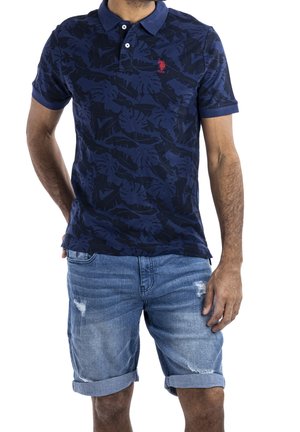 Homme portant un polo bleu marine à motif feuilles et un short en jean délavé avec revers, debout avec une main derrière le dos.