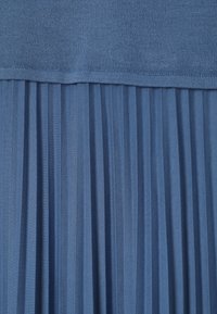 Sinine kangas, millel on ülaosas kootud osa ja alumises osas vertikaalselt volditud osa, näidates tekstuuri ja õmbluse üksikasju.