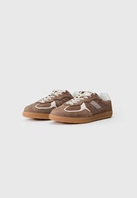 Zapatillas de ante marrón con detalles en blanco, perforaciones y una suela de goma. Los cordones son de color gris claro y cuentan con un collar acolchado.