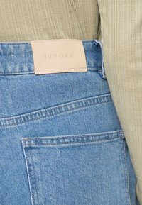 Lyseblå denimjeans med en lærreim merket "IVY OAK" på midjen, som viser tydelig søm og en teksturert ribbet topp.