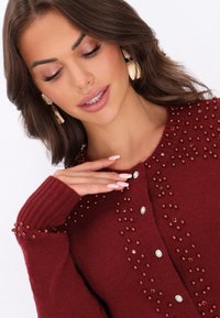 Burgundy stickad cardigan med ribbade manschetter, prydad med röda och klara pärlor längs framsidan och axlarna.
