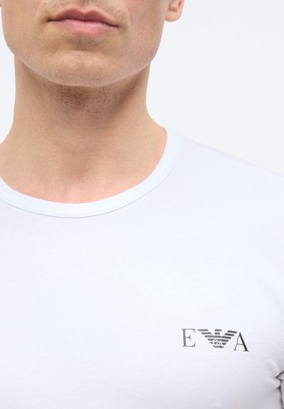 Κοντινό πλάνο ενός ατόμου που φοράει λευκό μπλουζάκι με στρογγυλή λαιμόκοψη και μικρό μαύρο λογότυπο Armani στη δεξιά πλευρά του στήθους.