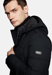 Czarna kurtka puchowa z kapturem, z teksturowanej tkaniny, zapinana na zamek, z naszywką z logo na rękawie. Gładkie wykończenie i ciepły design.