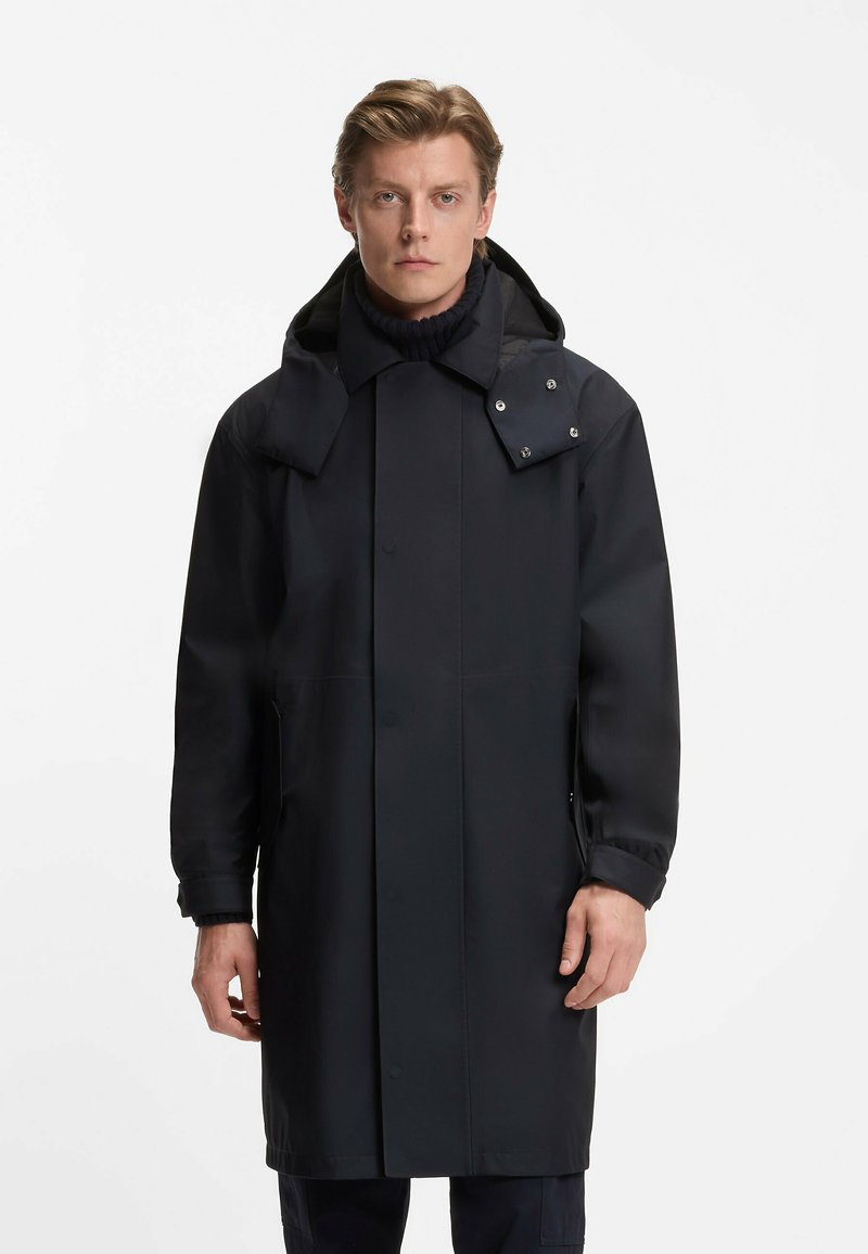 Cappotto lungo nero impermeabile con cappuccio, dotato di chiusure a pressione e tasche laterali. Il tessuto appare liscio e resistente all'acqua con una vestibilità sartoriale.