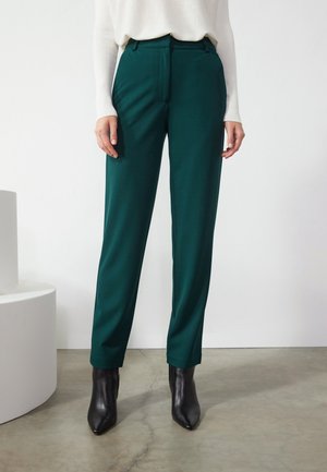 Femme portant un pantalon coupé sur mesure vert foncé, un haut blanc à manches longues et des bottines noires à bout pointu, debout sur un sol en béton.