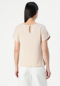 Top court beige clair à manches courtes avec un col rond et un détail en forme de clé à l'arrière avec une fermeture à bouton. Tissu lisse et design simple.