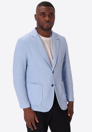 Uomo che indossa un blazer azzurro chiaro strutturato sopra una t-shirt bianca e pantaloni neri, in piedi davanti a uno sfondo uniforme.
