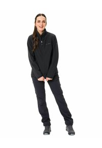 Giacca softshell nera con zip, dotata di un colletto alto, abbinata a pantaloni scuri e robusti scarponi da trekking grigi, su uno sfondo bianco.