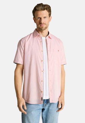 Mann trägt ein hellrosa kurzärmeliges Hemd über weißem T-Shirt und hellblauen Jeans, steht vor schlichtem Hintergrund.