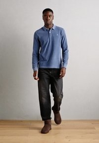 Polo shirt à manches longues bleu clair, jean foncé et bottines marron. Design simple avec un petit logo sur la poitrine. Texture lisse.