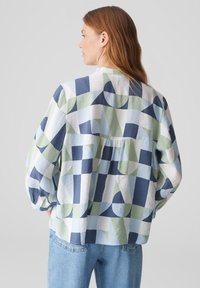 Camicia ampia multicolore con motivi geometrici in blu navy, verde e azzurro; presenta maniche lunghe e un dettaglio plissettato sul retro.