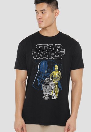 Man wearing black Star Wars t-shirt with Darth Vader, R2-D2, and C-3PO graphics looking to the side against a plain light background.