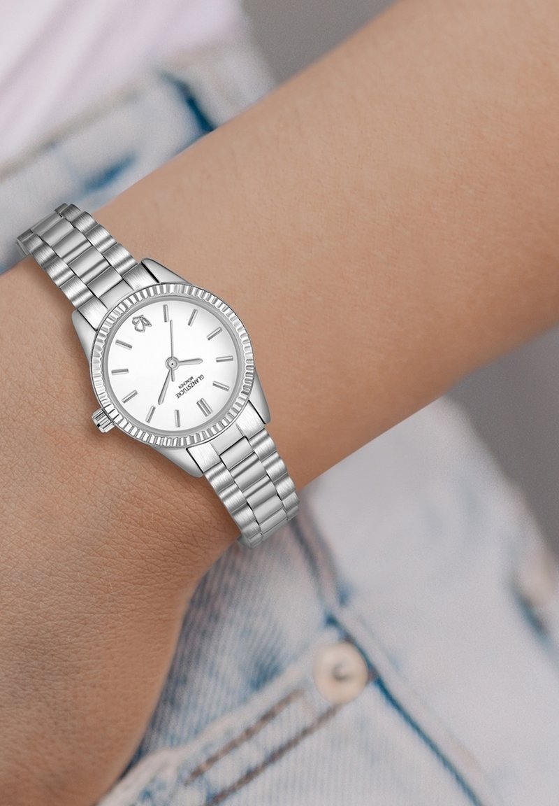 Hõbedane metallist käekell ümmarguse valge dialli, tunni markide ja volditud äärisega. Omab lingitud käeosa ja poleeritud viimistlust.