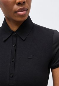 Κοντινό πλάνο ενός ατόμου που φοράει μαύρο πουκάμισο με γιακά και κουμπιά, με ένα διακριτικό κεντητό λογότυπο στο αριστερό στήθος.