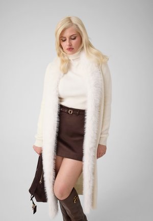Femme blonde portant un long manteau blanc bordé de fourrure, un col roulé blanc, une jupe marron et des bottes marron jusqu'aux genoux, tenant un sac à main marron.
