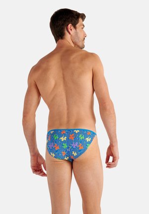 Man facing away wearing blue swim briefs with multicolored floral patterns on a plain light background.