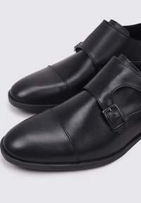 Zapatos de cuero negro con una textura suave, punta en forma de almendra, doble correa y detalle de hebilla, con una suela de goma de perfil bajo.