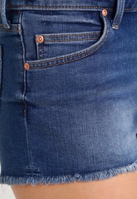 Pantalones cortos de mezclilla en azul oscuro con un dobladillo deshilachado, que cuentan con dos bolsillos frontales y herrajes metálicos en tono cobre. Tela suave y texturada.