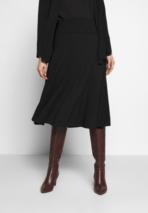 Vrouw in een zwarte rok tot halverwege de knie en bruine leren laarzen met hakken tot over de knie, staand tegen een effen witte achtergrond.