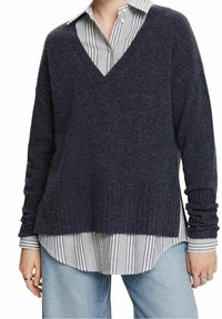 Navy V-Ausschnitt-Pullover aus weichem Strickmaterial, mit geripptem Saum und Bündchen, über einem hellblauen und weißen gestreiften Hemd getragen.