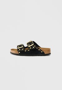 Sandale à glissière en suede noir avec deux sangles ajustables en or, des clous décoratifs dorés et une semelle en liège texturée.