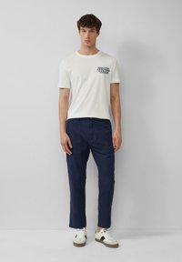 Witte T-shirt met ronde hals en grafische print op de linkerborst, gecombineerd met marineblauwe broek. Model draagt witte en zwarte sneakers.