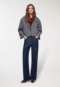 Blazer grigio in lana cropped con maniche oversize, indossato sopra un top borgogna e jeans a gamba larga blu scuro. Scarpe a punta rosso scuro.