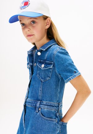 Niña joven con cabello largo y rubio, vestida con un vestido azul de mezclilla y una gorra blanca con visera azul, de pie con las manos en los bolsillos contra un fondo blanco.
