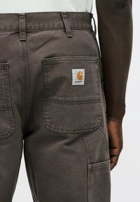 Σκούρο καφέ παντελόνι εργασίας με τσέπες κεντήματος, λογότυπο Carhartt στην δεξιά πίσω τσέπη, φορεμένο από άτομο με σκούρο δέρμα και σκούρο μακρυμάνικο πουκάμισο.