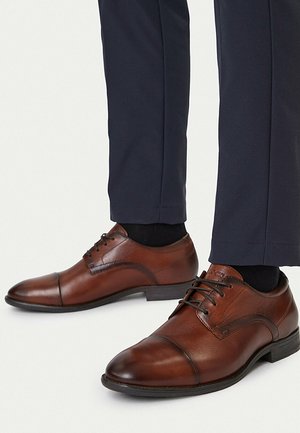 Braune Lederschuhe mit schwarzen Schnürsenkeln, getragen zu dunkelblauen Hosen und schwarzen Socken, vor weißem Hintergrund.