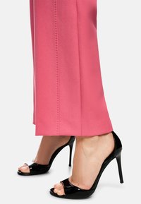 Schwarze offene High Heels mit glänzender Oberfläche, kombiniert mit rosa maßgeschneiderten Hosen, die seitliche Nähte und eine glatte Textur aufweisen.