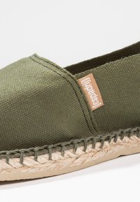 Green canvas espadrille featuring a beige woven jute sole, contrasting stitching, and a small branded tag on the side.