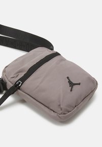 Gray nylon crossbody bag with a black zip, adjustable strap, and a black Jordan logo on the front. Compact, rectangular design.