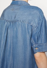 Widok z tyłu osoby noszącej jasnoniebieską koszulę dżinsową z krótkim rękawem i kołnierzykiem, widoczne przeszycia i detale z guzikami na mankiecie rękawa.