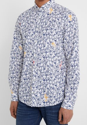Mann trägt ein weißes Hemd mit Knöpfen und blauen, roten sowie orangefarbenen Flamingo-Mustern, kombiniert mit blauen Jeans, steht vor einem einfarbigen Hintergrund.
