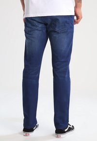 Pantalones vaqueros de mezclilla azul oscuro con un diseño de pierna recta, que cuentan con bolsillos traseros y un ligero desgaste. Combinados con zapatillas negras y blancas.