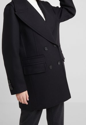 Manteau noir croisé à double boutonnage avec larges revers et poches à rabat, porté sur un haut blanc et un pantalon foncé, bras levé partiellement visible.