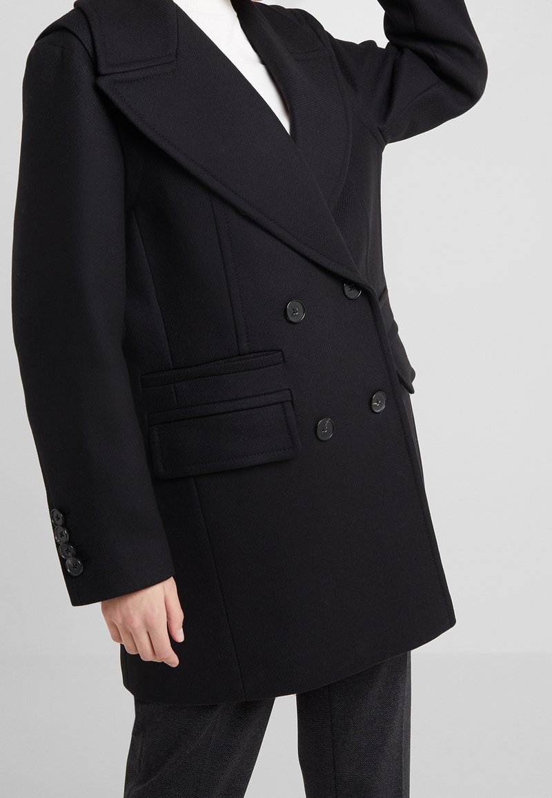 Manteau noir croisé à double boutonnage avec larges revers et poches à rabat, porté sur un haut blanc et un pantalon foncé, bras levé partiellement visible.