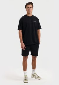 Zwarte t-shirt met klein wit logo, gecombineerd met zwarte shorts. Casual fit, korte mouwen, gemaakt van zachte stof, gedragen met lichte sneakers.