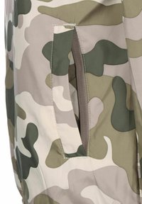 Rohelise ja beeži toonides kamuflaažimustriga kangas, millel on tugevdatud õmblustega tasku detail. Sile, matt pind.