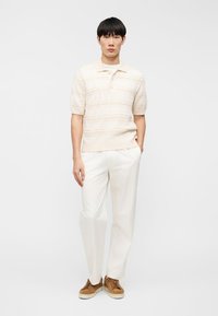 Νεαρός άντρας στέκεται με το ένα χέρι στην τσέπη, φορώντας ελαφριά πλεκτή μπλούζα πόλο σε κρεμ απόχρωση, λευκό παντελόνι και καφέ σουέντ αθλητικά παπούτσια σε απλό φόντο.